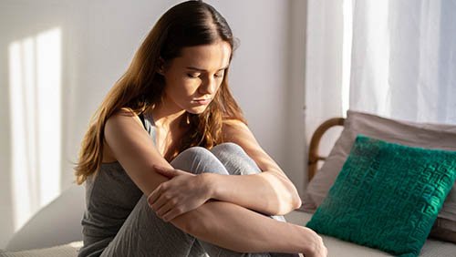 Женщина на кровати в светлой комнате, обнимает колени.